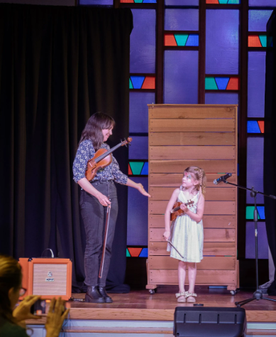 Woman on stage with a violin gestures to a child with a violin, wooden backdrop.