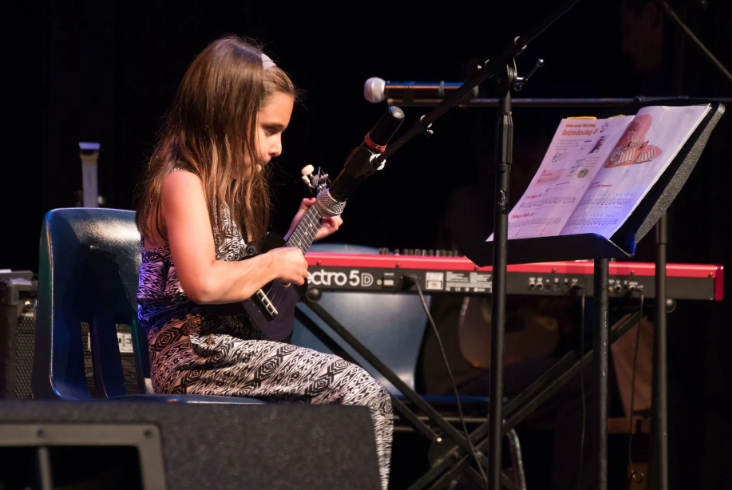 Young girl playing ukulele on stage with keyboard and sheet music.