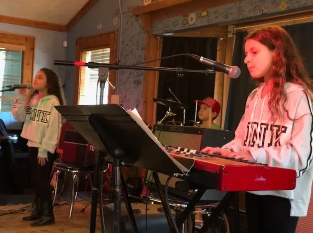Two girls singing and playing keyboard on stage. A boy watches from behind drums.