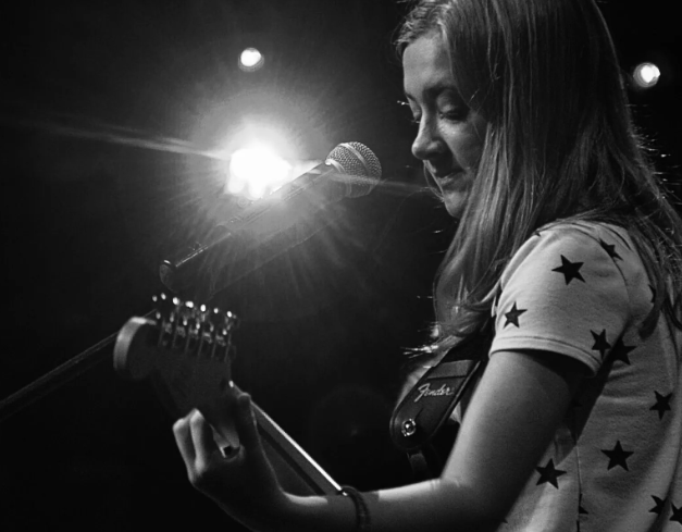 Woman playing electric guitar, lit by stage light, microphone in front.