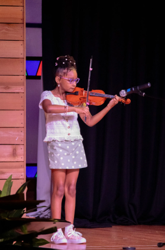 Young person playing violin on stage; wearing glasses, polka dot skirt, white top.