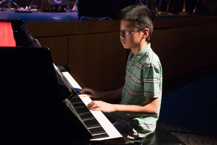 Boy with glasses playing a grand piano on a stage, lit with stage lighting.