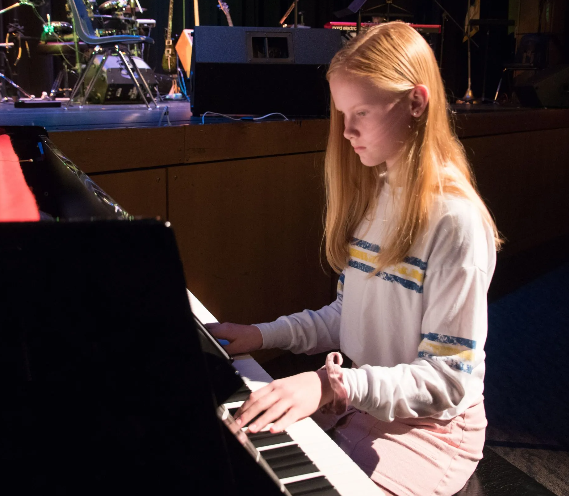 Young person with long red hair plays piano on stage.