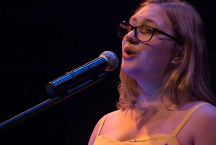 Woman singing into a microphone on stage, wearing glasses and a yellow top; dark background.