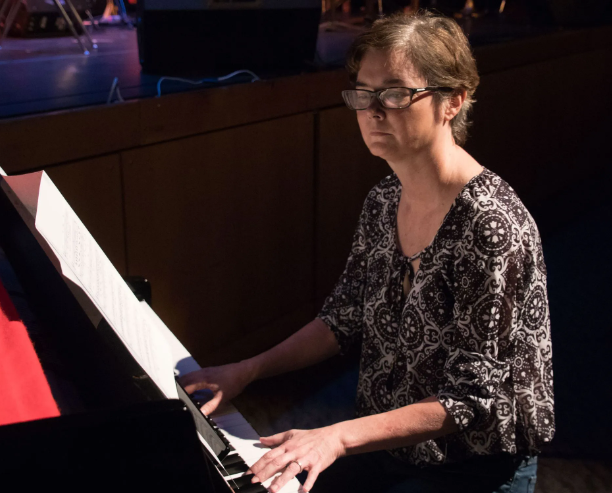 Woman playing a piano, wearing glasses and patterned shirt. Performing indoors, lit by stage lights.