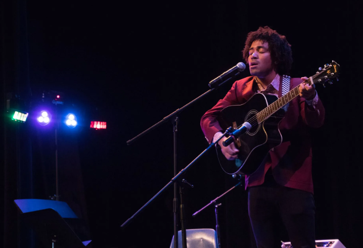 Person with curly hair singing and playing an acoustic guitar on stage with a microphone.