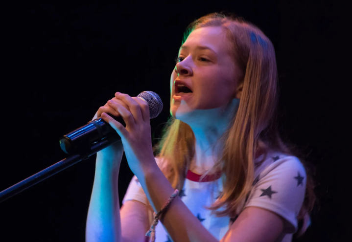 Young person singing into a microphone on stage, mouth open, looking up, under stage lights.
