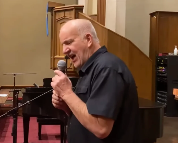 Man singing into a microphone in a church. He wears a black shirt and looks focused. A piano is behind him.