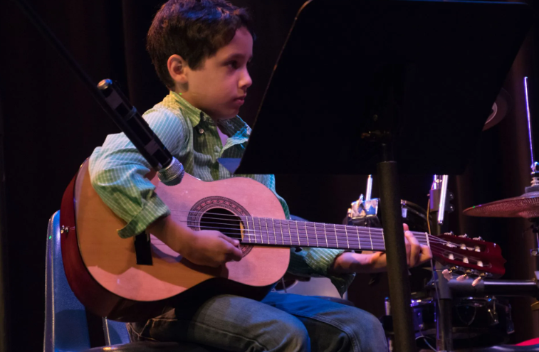 Young child playing an acoustic guitar on a stage, focused expression. Microphone nearby.