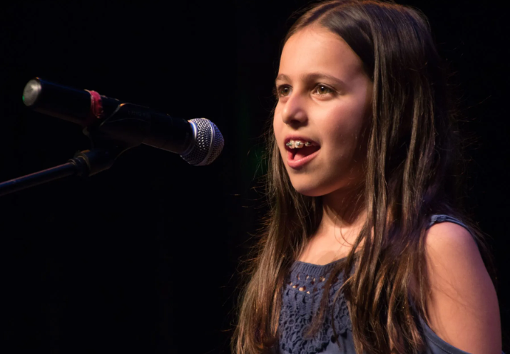 Young person with braces singing into a microphone on a stage with a dark background.