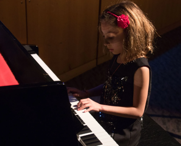 Young girl with a red hair accessory plays a black piano in a dim-lit space.