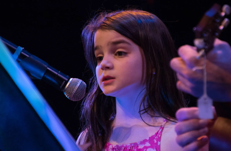 Girl singing into a microphone onstage; pink dress, long dark hair; person holding a guitar.