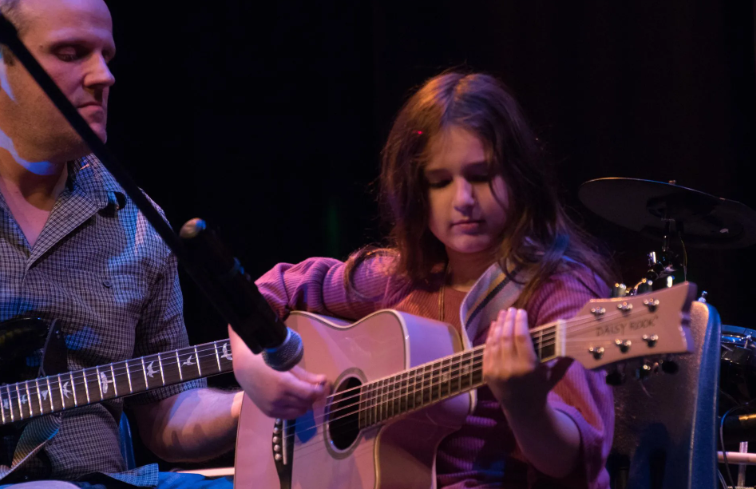 Girl playing acoustic guitar on stage, next to a man playing electric guitar.