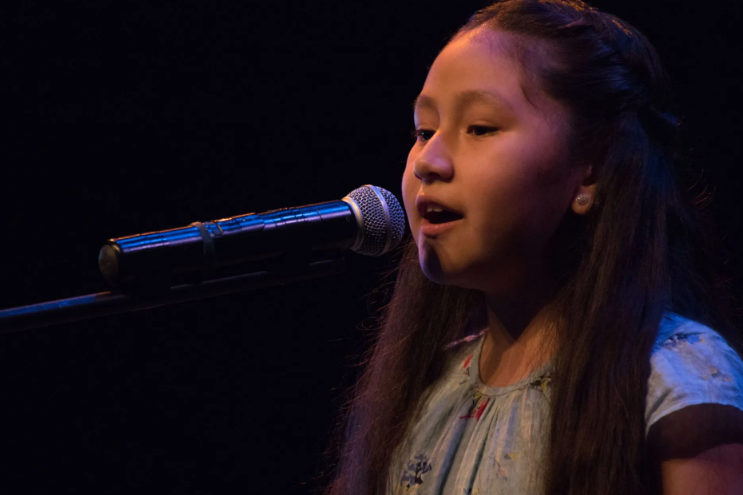 Young person singing into a microphone on a dark stage.