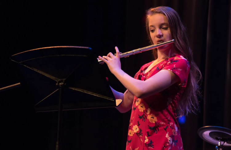 Woman in red floral dress playing a silver flute on stage, music stand in front.