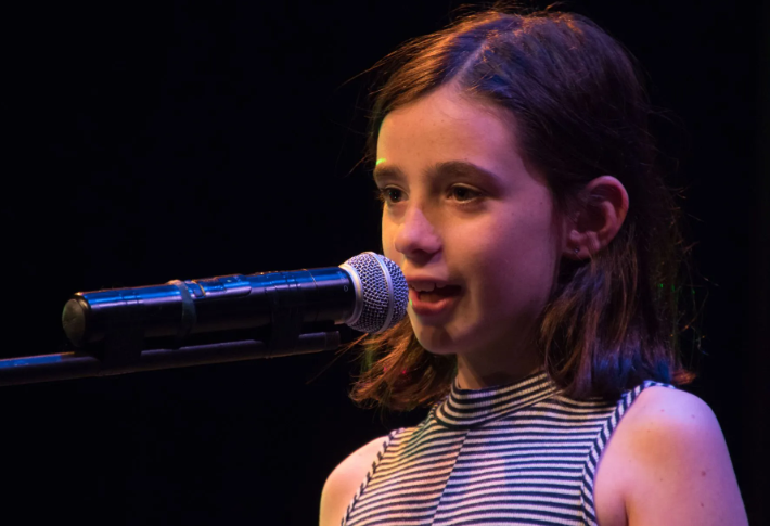 Girl singing into a microphone on stage, wearing striped top. Dark background.