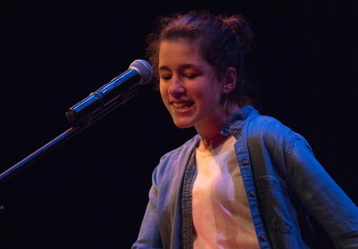 Young person singing into a microphone on stage, wearing a blue shirt and white undershirt.