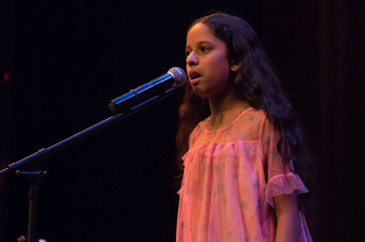 Girl singing into a microphone on stage, wearing a pink dress, dark hair.