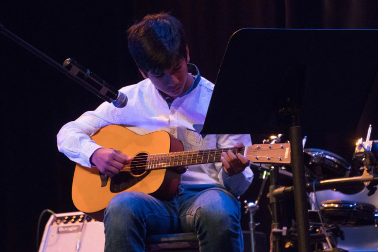 Person playing acoustic guitar on stage, reading sheet music.