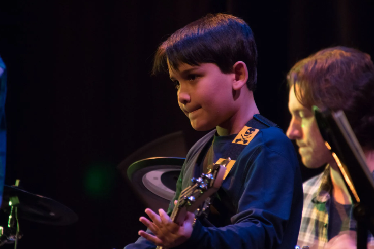 Boy with a guitar plays on stage. He wears a blue shirt and has a focused expression. A person is beside him.