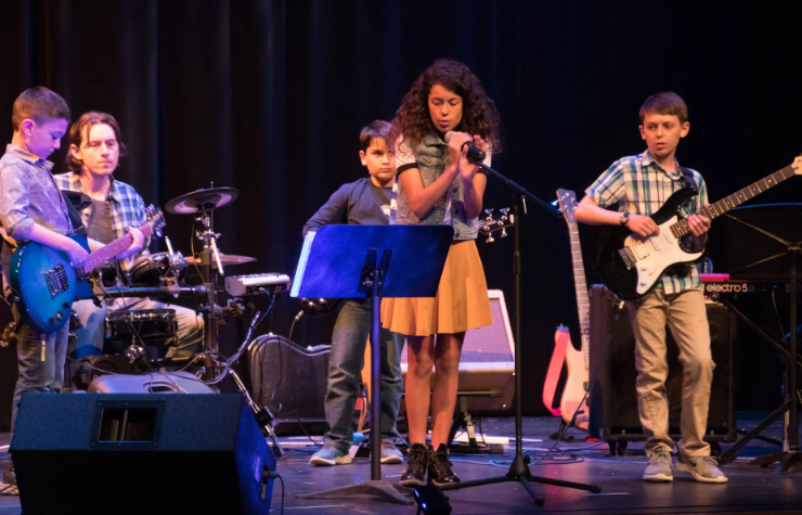 A young band performs on stage: a singer, guitarists, drummer, and bass player.
