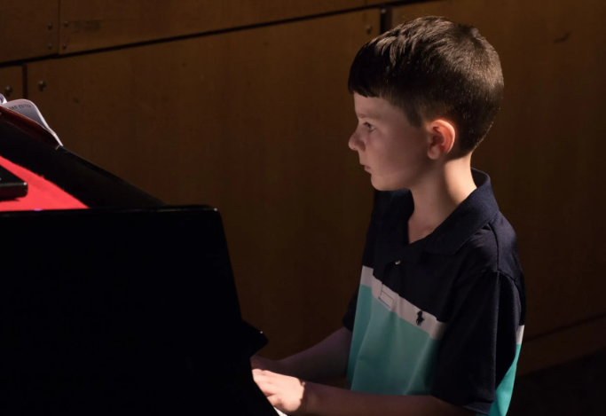 Boy playing piano, lit from the side. Wearing a polo shirt, looking at sheet music. Dark setting.