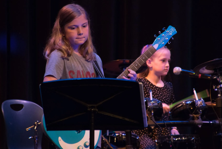 Two young girls perform music on stage. One plays guitar, the other drums.