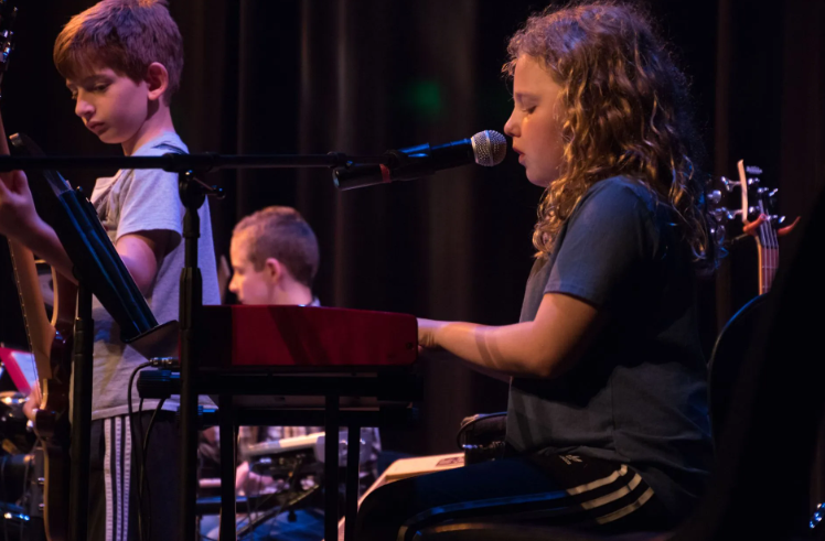 Girl singing and playing keyboard on stage with bandmates. Low light, dark background.
