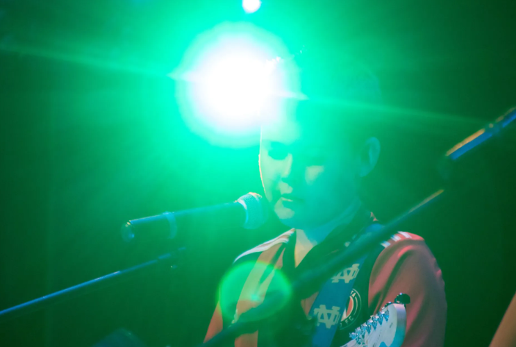 Person singing and playing guitar on stage, lit with bright green light.
