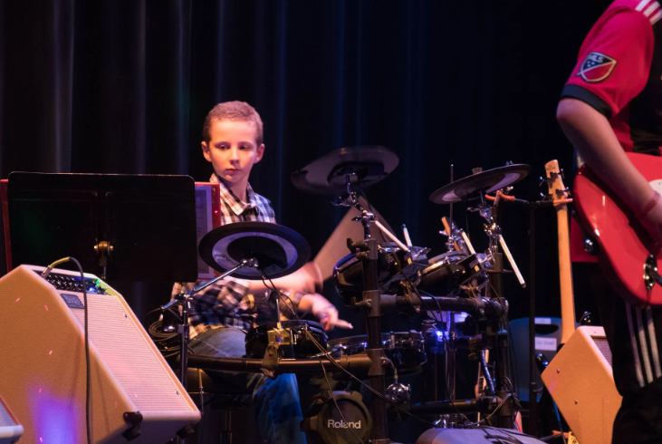 Boy playing an electronic drum set on a stage with a band, dark background, focused expression.