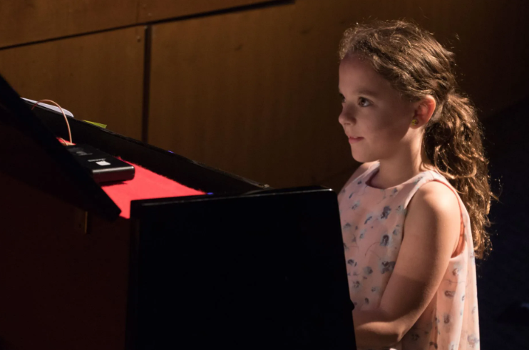 Young girl with curly hair stands by a piano, illuminated by stage lighting, looking to the side.