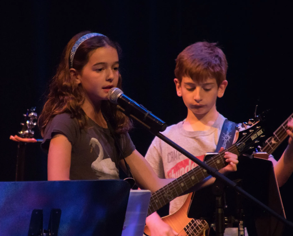 Girl singing into a microphone, boy playing guitar on stage. Dark setting, blue lighting.