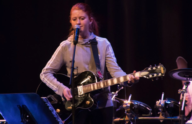 Woman with red hair playing a black guitar and singing into a microphone on stage.