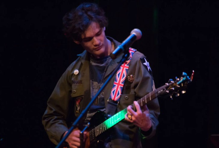 A person playing an electric guitar onstage under dim lighting. They wear a jacket and a Union Jack strap.