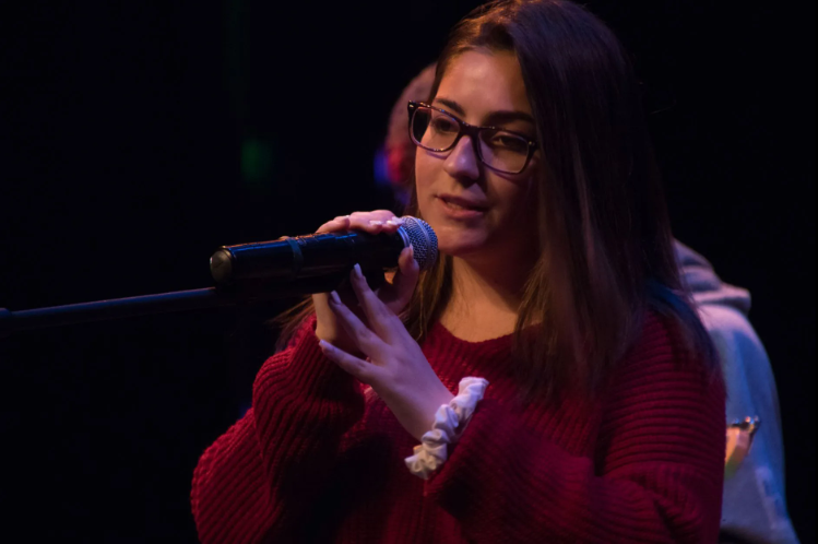 Woman with glasses singing into a microphone on stage, wearing a red sweater.