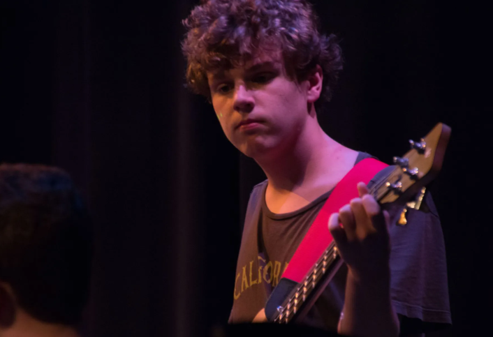 Person with curly hair, holding a bass guitar with a red strap, on stage in front of a dark backdrop.