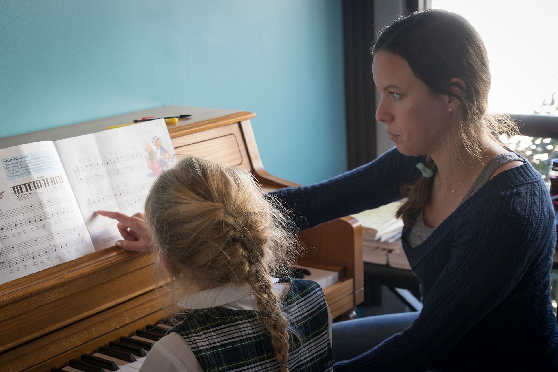 Woman points at sheet music for child at a piano, indoors.