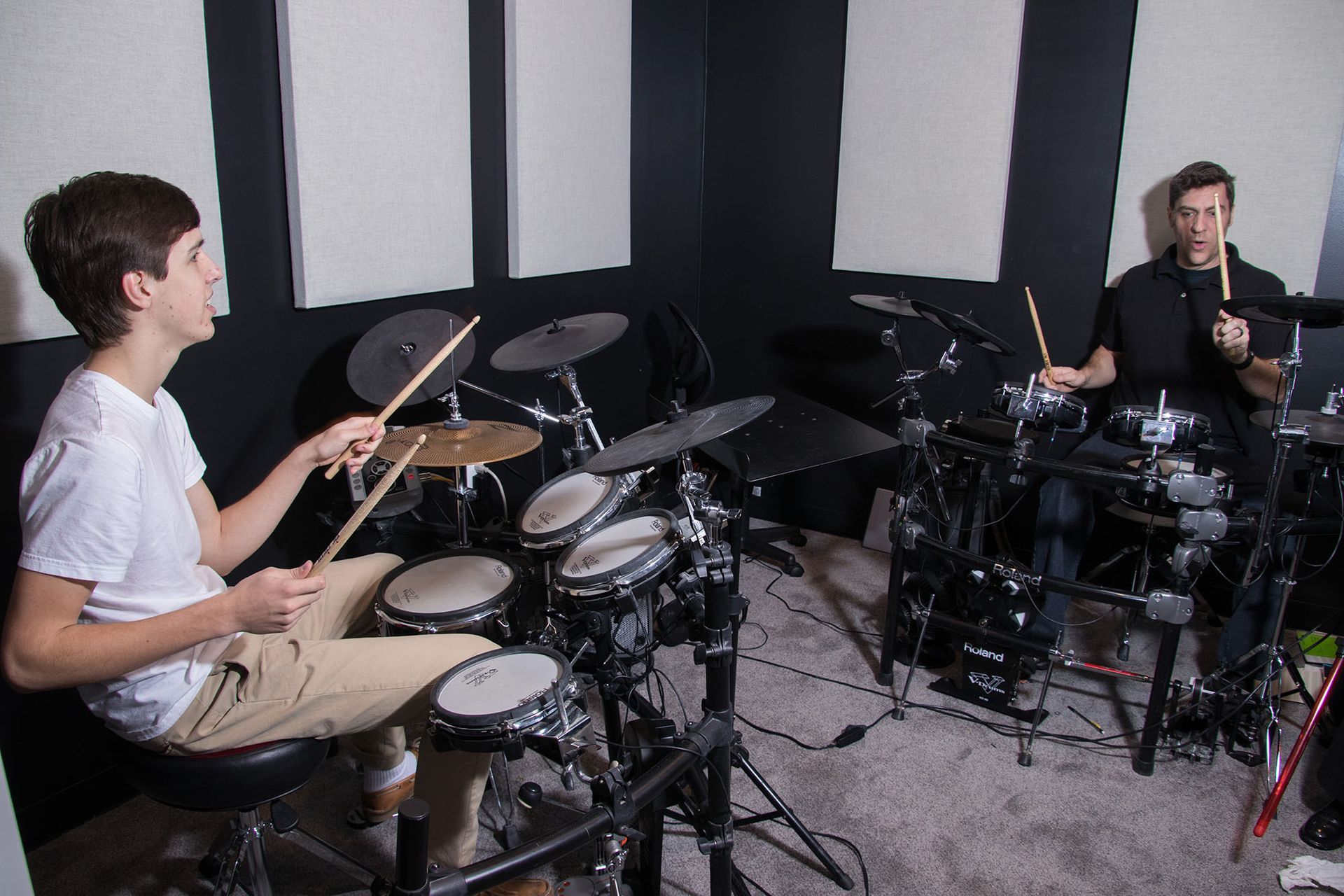 Two people playing electronic drums in a black-walled studio, one on the left in white, the other on the right in black.