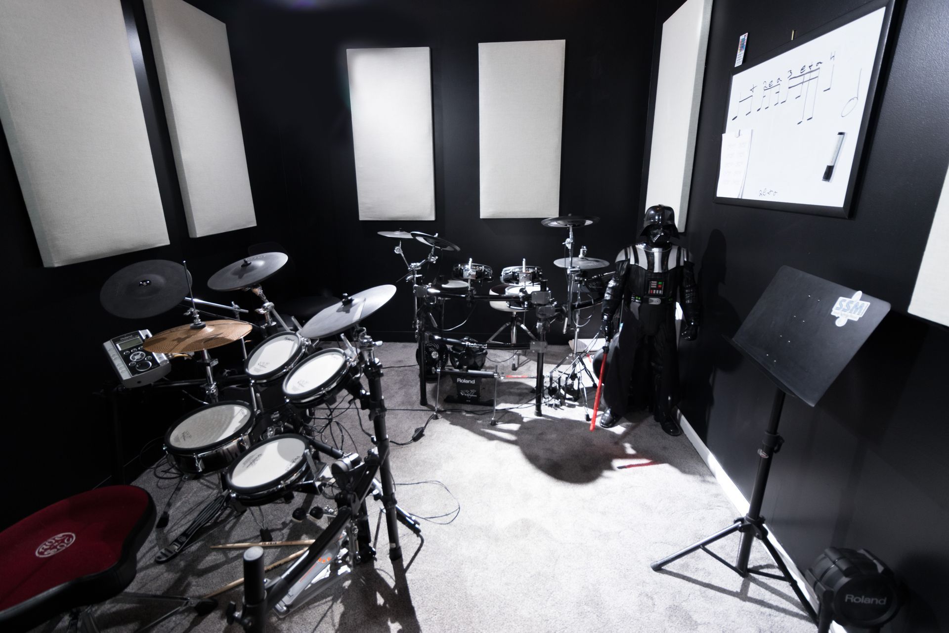 A black room with an electronic drum set, acoustic panels, music stand, and Darth Vader statue.