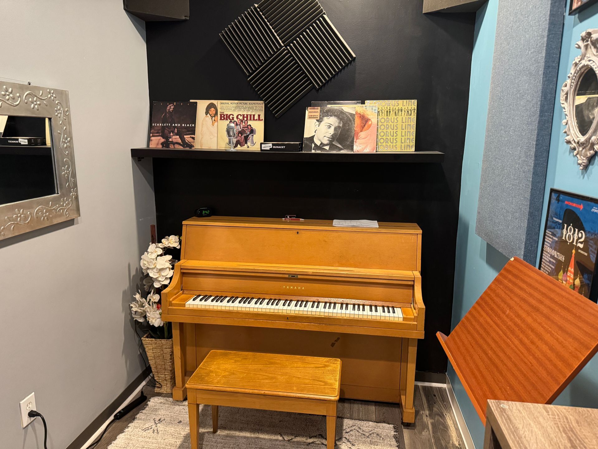 Piano in a room with a shelf holding records. A wooden stool is in front. The walls are gray, black, and blue.