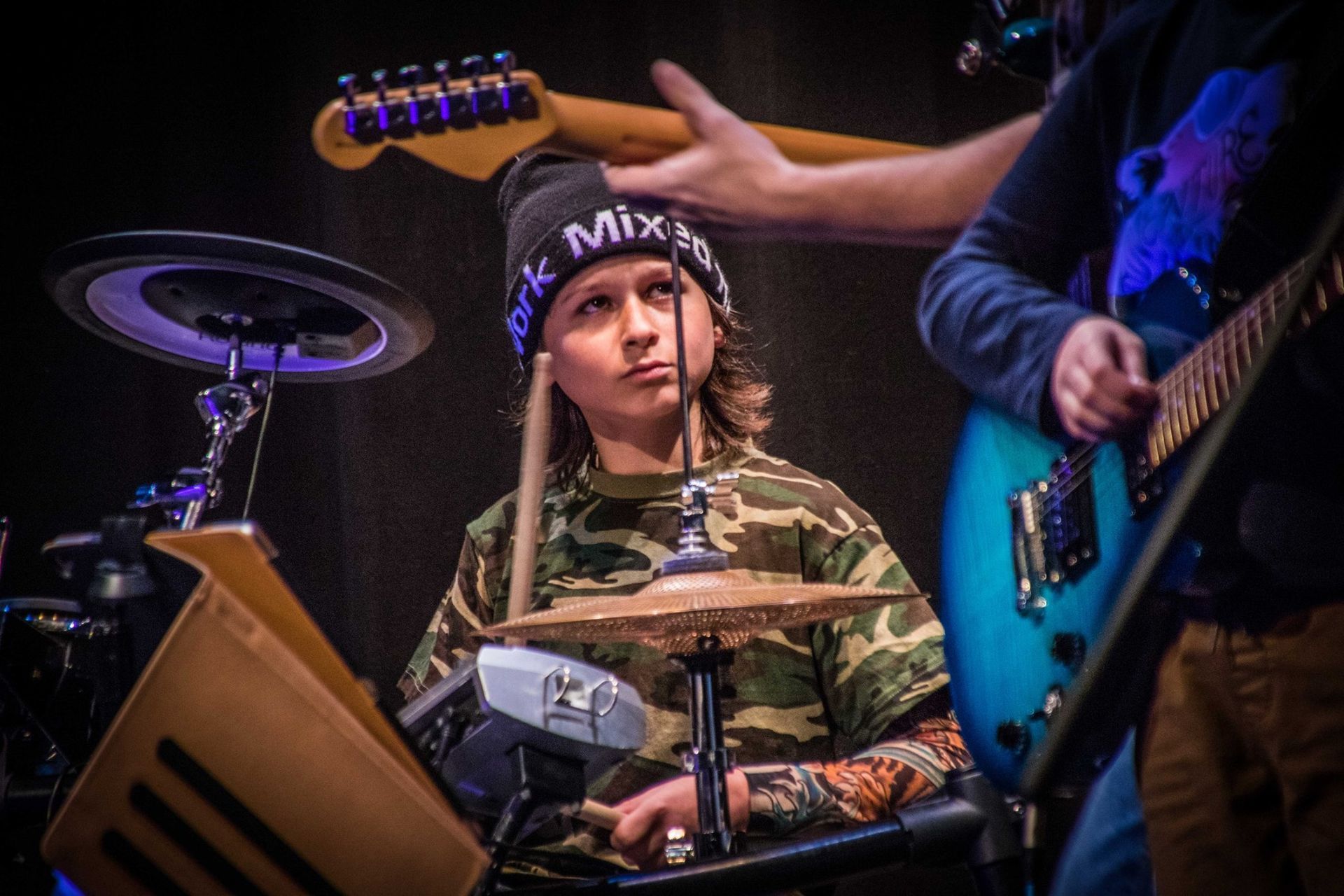 A drummer in camouflage shirt and black beanie performs. Other musicians with guitars are partially visible. Stage setting.