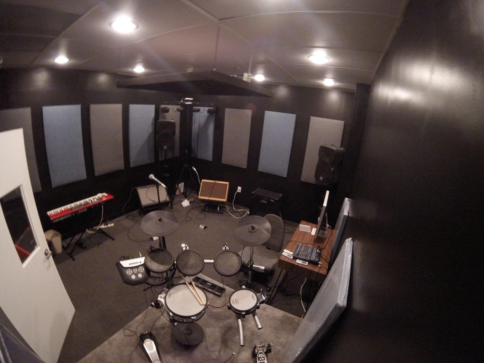 Music recording studio with drums, keyboard, and soundproofing panels. Dark walls, overhead lights.
