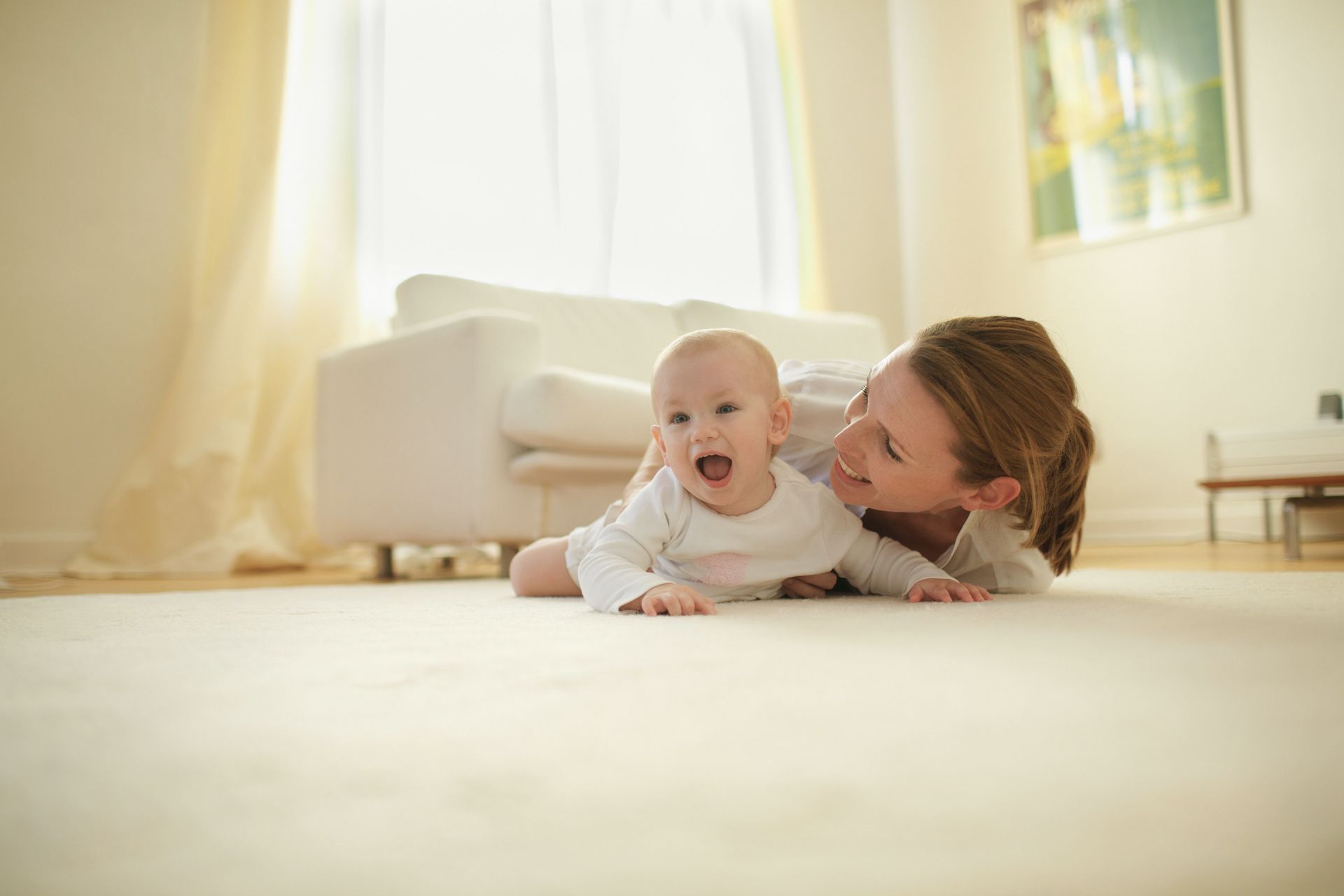 Woman and baby on floor, both smiling. Bright room with white furniture and carpet.