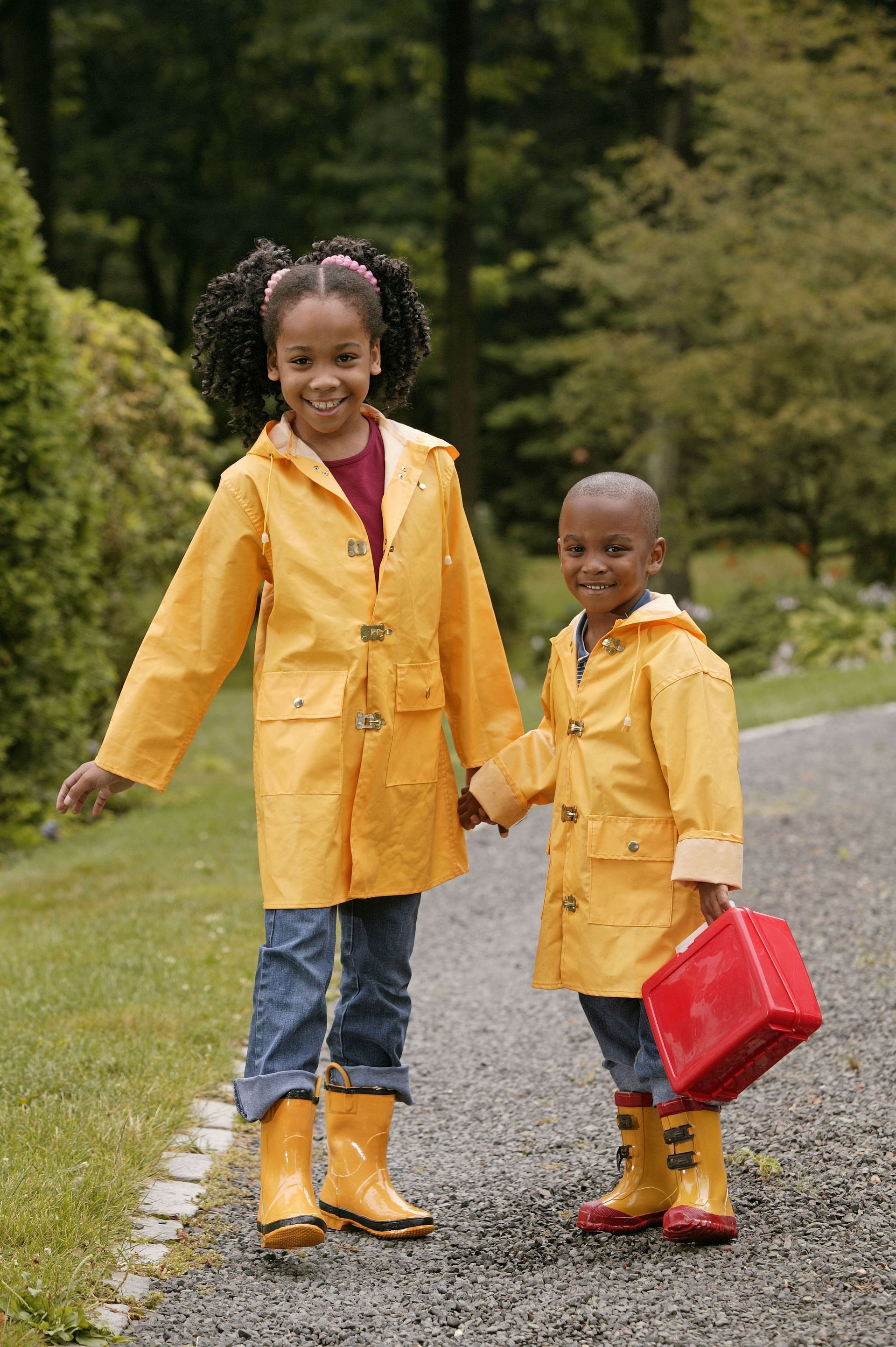 Two children in yellow raincoats and boots hold hands on a stone path.