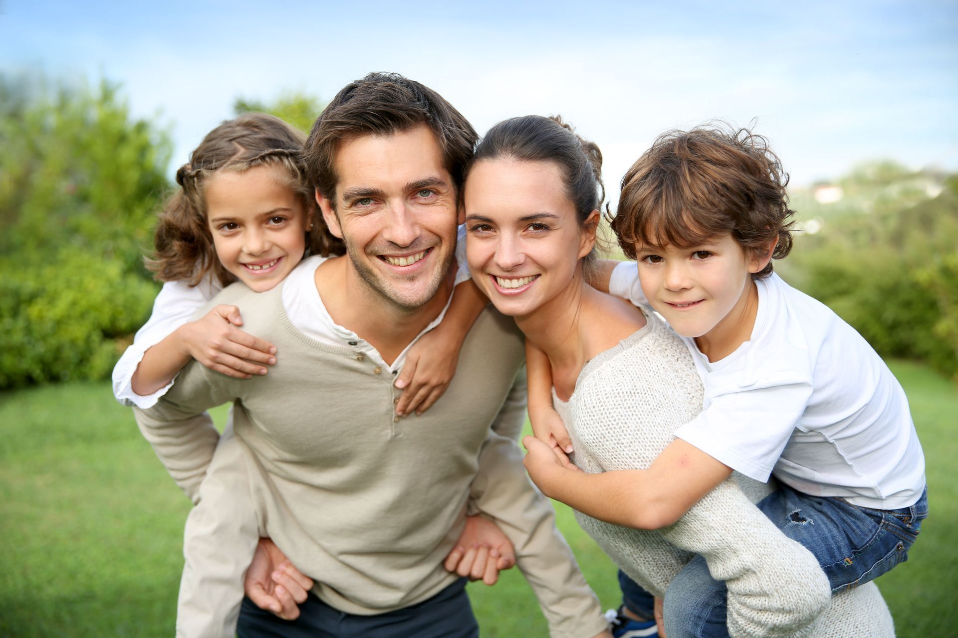 Family of four smiling, piggybacking in a sunny outdoor setting with green grass.