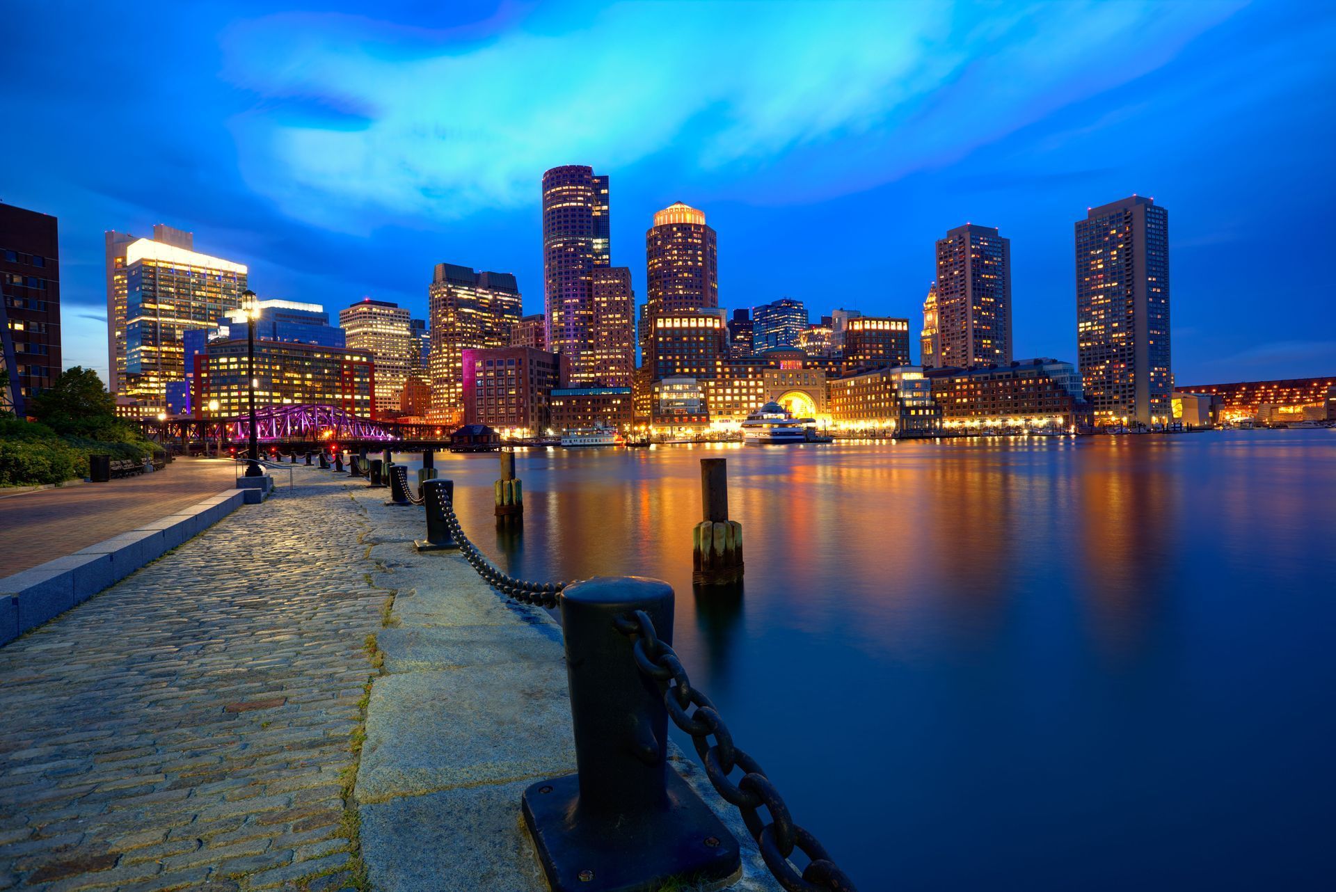 Boston skyline at dusk, illuminated buildings reflected in calm water.