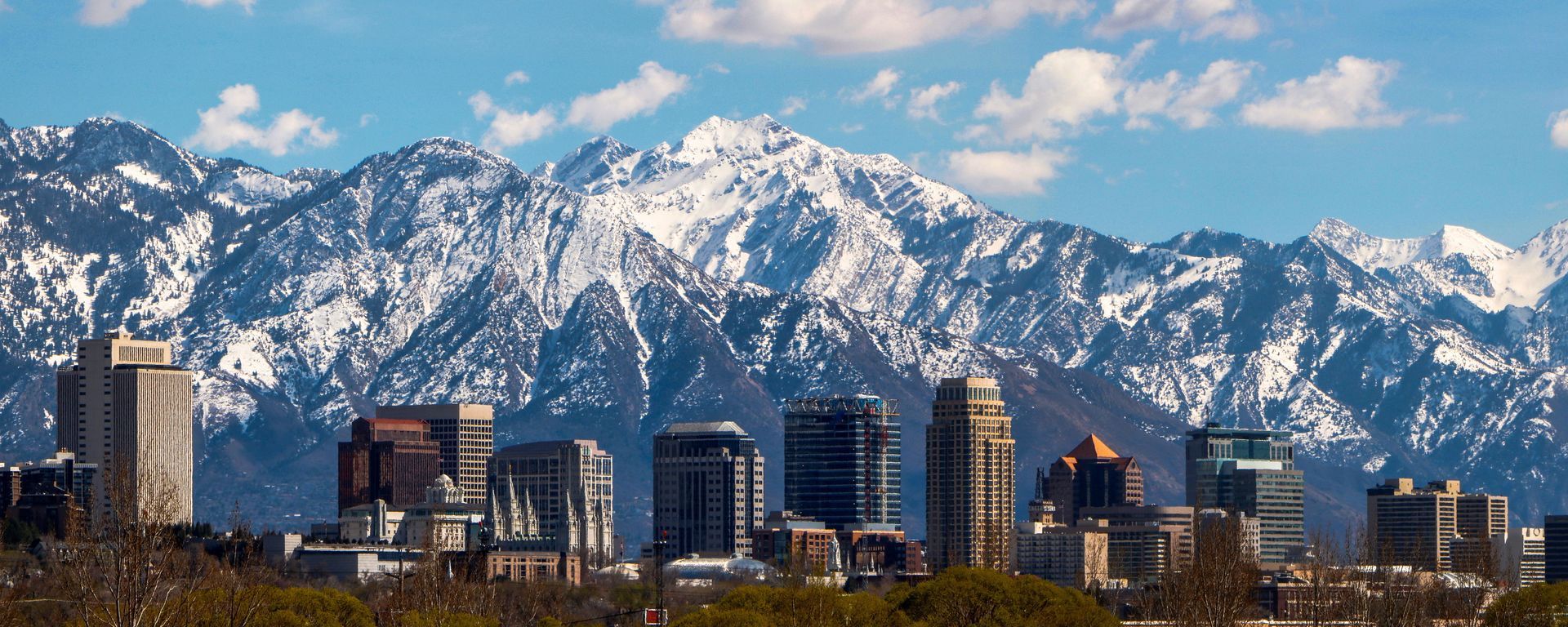 Snow-covered mountains rise behind a city skyline under a clear, blue sky.