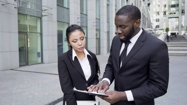 Man and woman in suits reviewing documents outside a modern building.