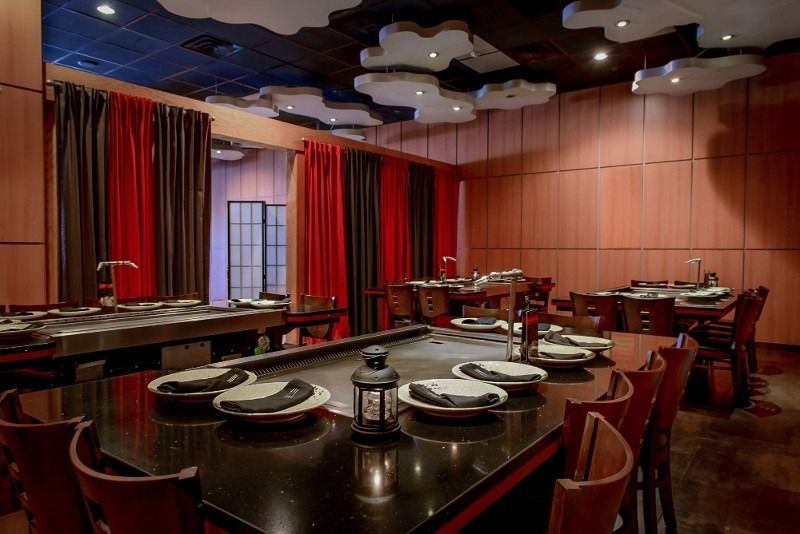 Teppanyaki restaurant interior with cooking surfaces, tables, chairs, and cloud-like ceiling decorations. Red and brown curtains.