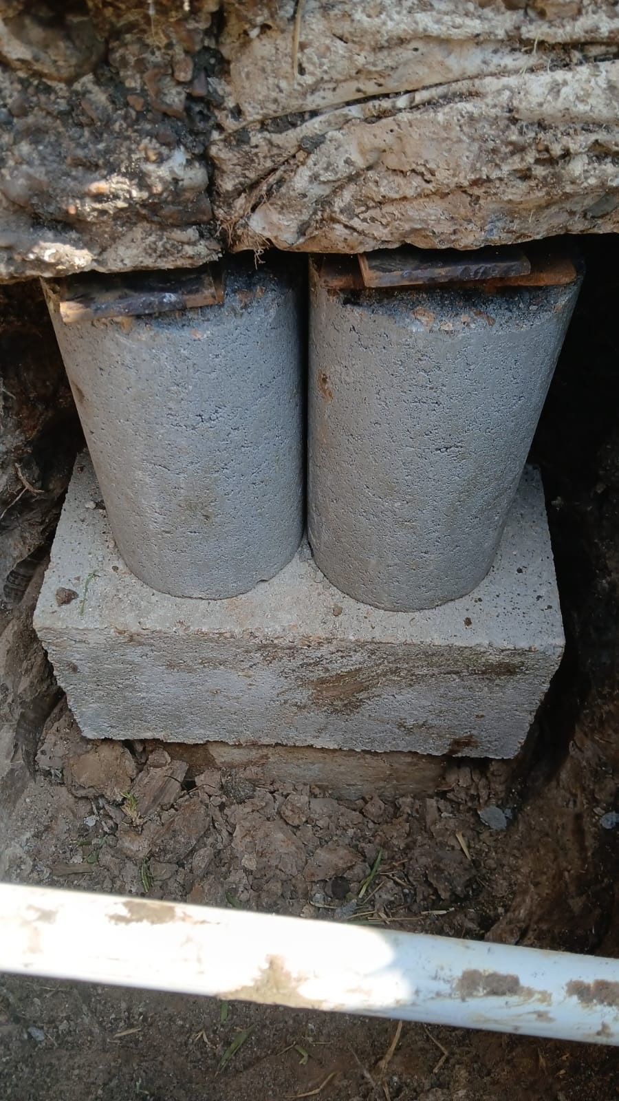 Two gray concrete columns support wooden structure, sitting on rectangular concrete base in dirt